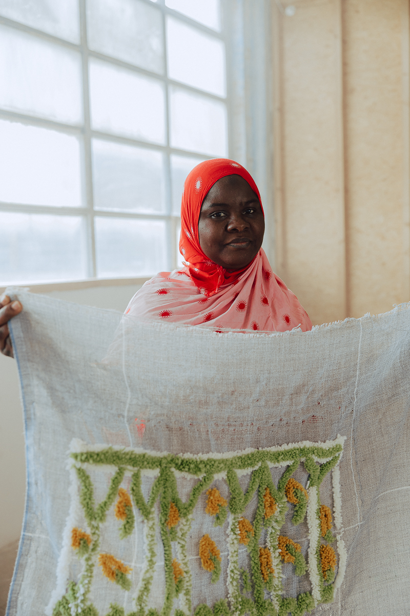 Une femme noire qui porte un foulard rouge tient dans ses deux mains une pièce textile tuftée blanche verte et jaune. Une lumière chaleureuse provient de la fenêtre à l'arrière de la femme.